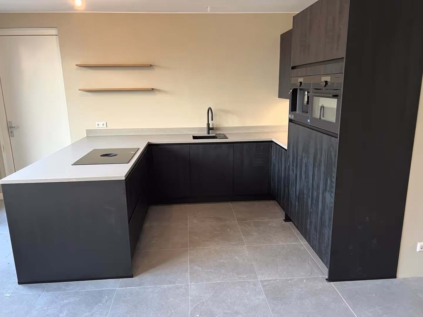 A modern black kitchen and dining area with white floors, a round dining table with chairs, and a geometrically-designed chandelier. There's also a fireplace with stacked logs on the right